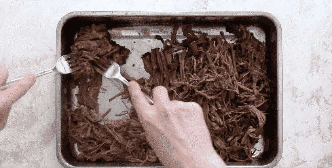 Shredding the beef with a fork in a silver pan. 