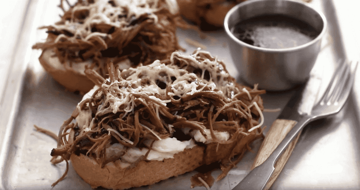 Roast beef sandwich with mashed potatoes, gruyere cheese and au jus on a platter with a fork and knife.