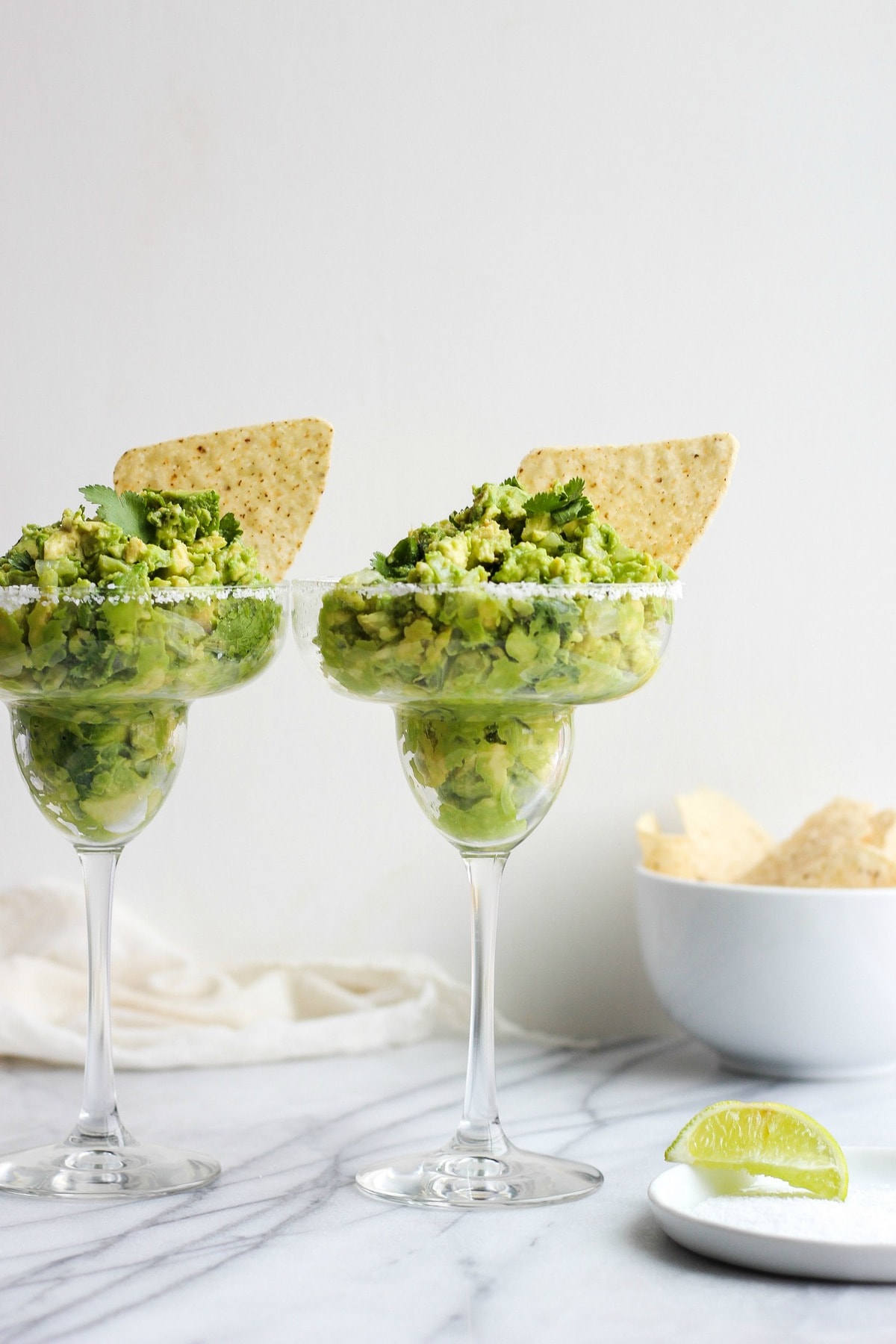 two margarita glasses filled with tequila guacamole, next to a small bowl of tortilla chips