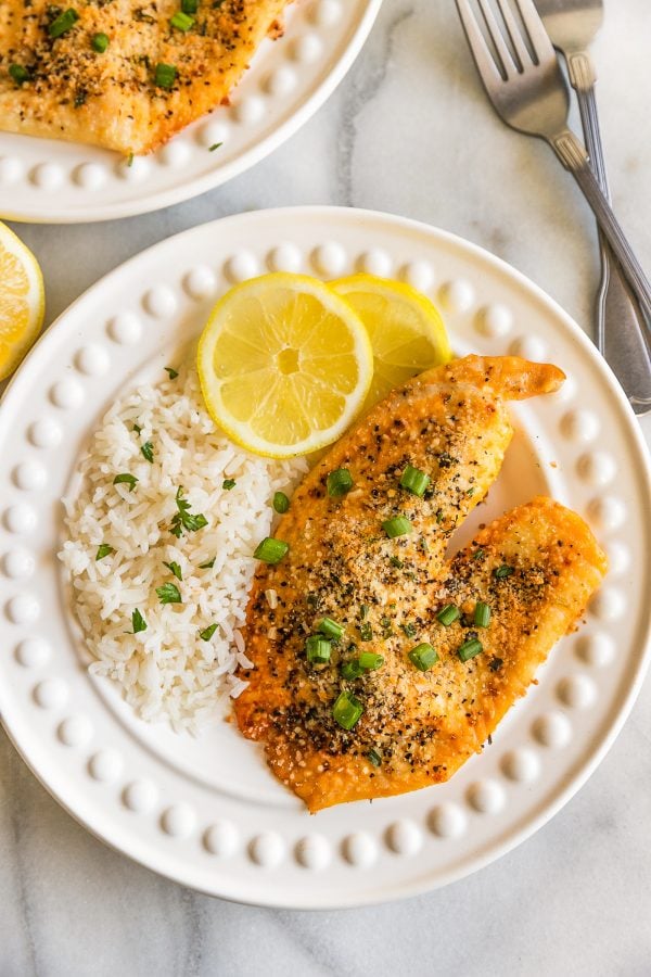 parmesan crusted tilapia on a white dinner plate, garnished with white rice, lemon slices, and green onions