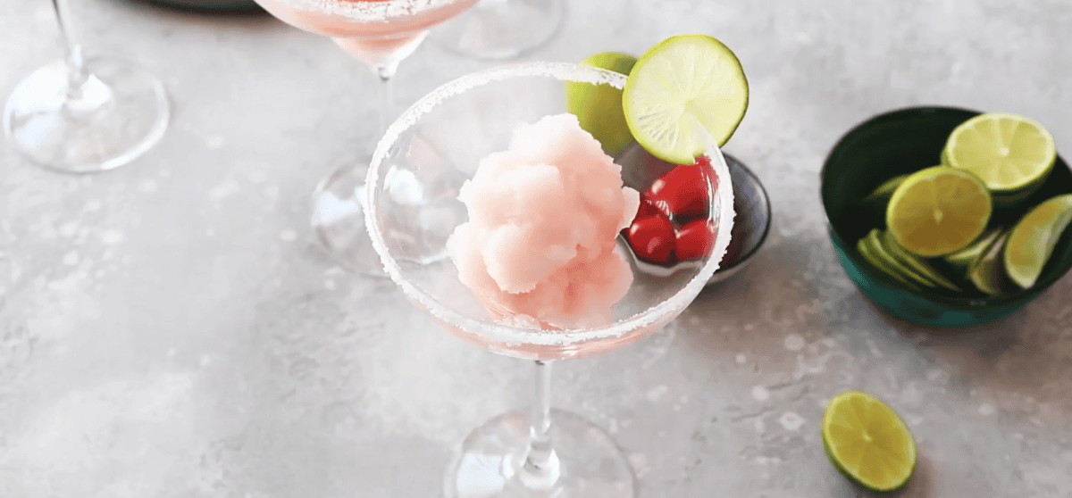Frozen cherry limeade margarita in a salt-rimmed glass with a lime with a bowl of limes and a bowl of cherries in the background