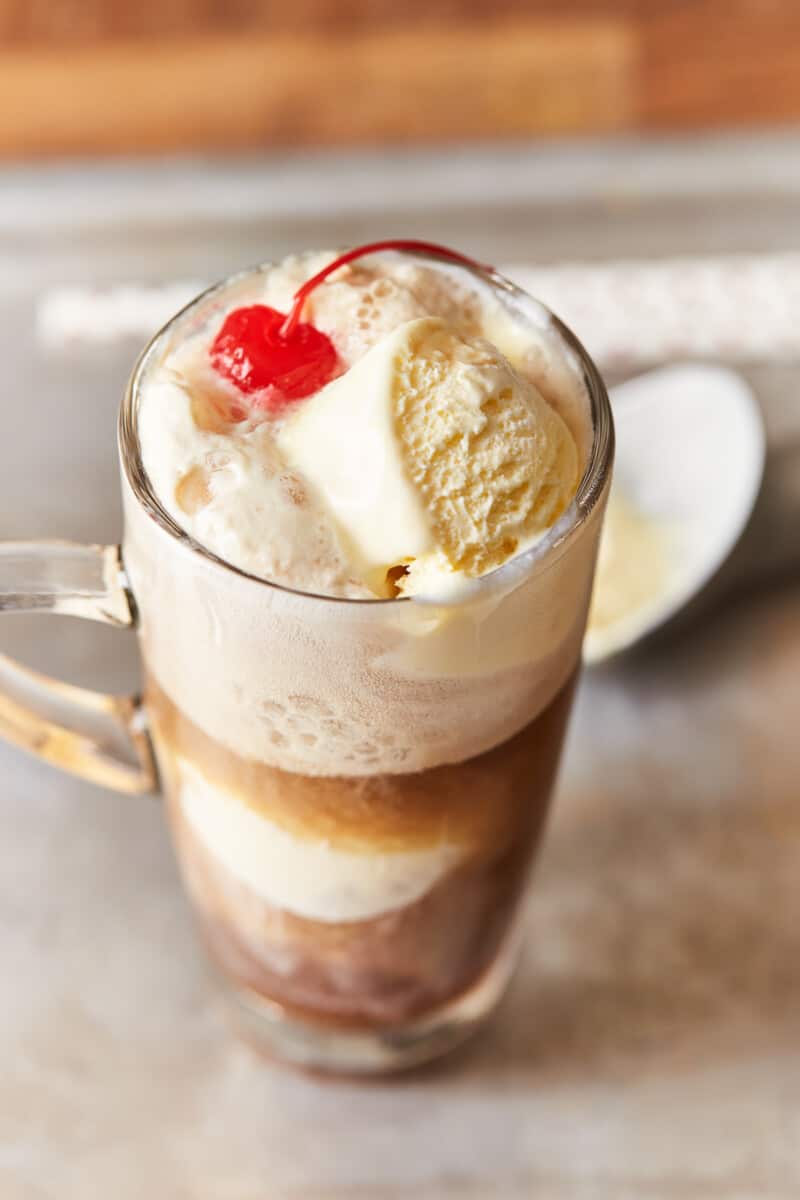 three-quarters view of an alcoholic root beer float in a tall clear glass with a handle with a cherry on top.