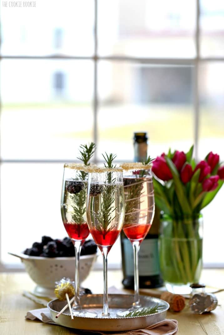 Cocktails on a table with champagne, bowl of blackberries, and a vase of tulips in front of a vindow
