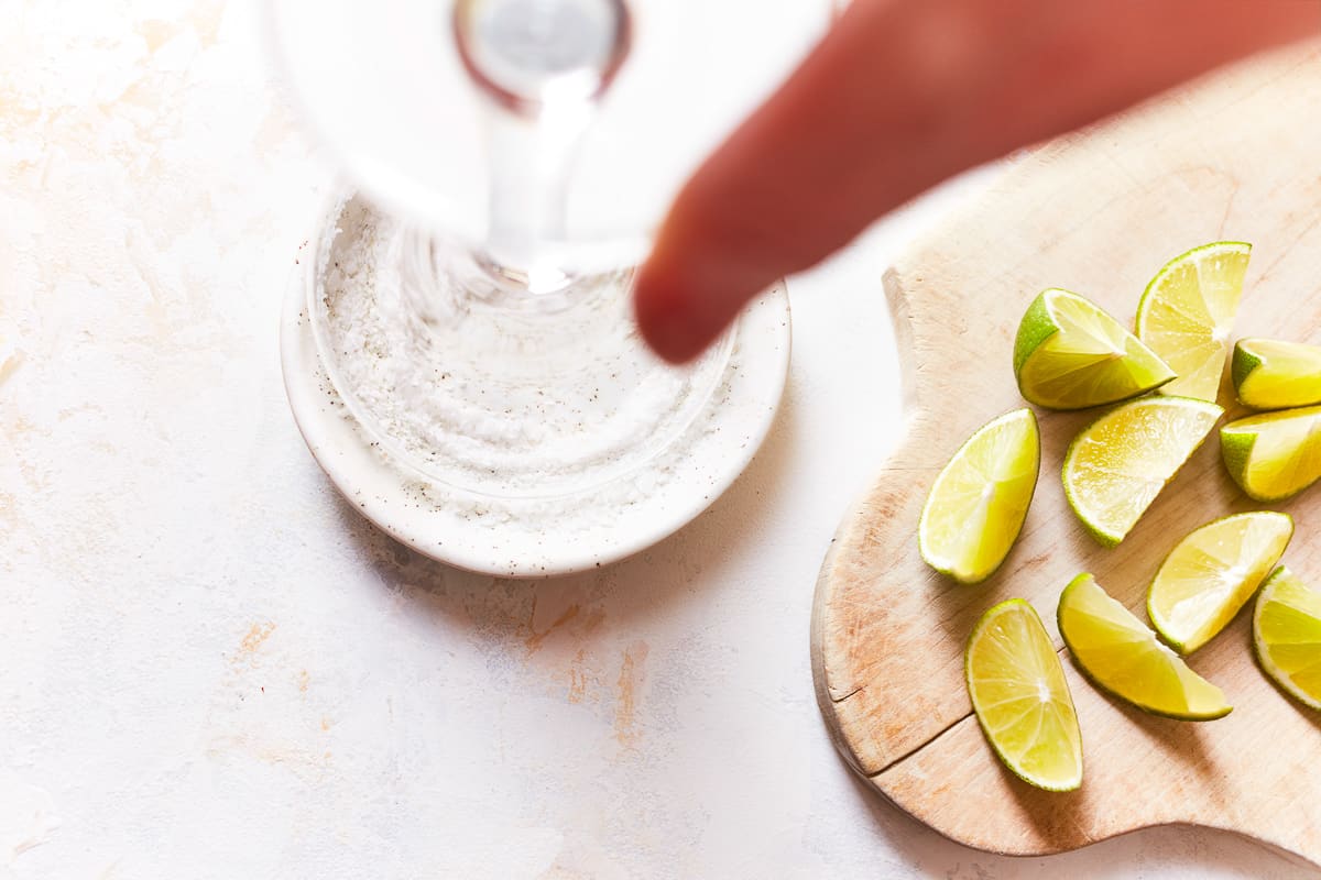 A hand dipping the rim of a glass into salt and lime wedges on a wooden cutting board.