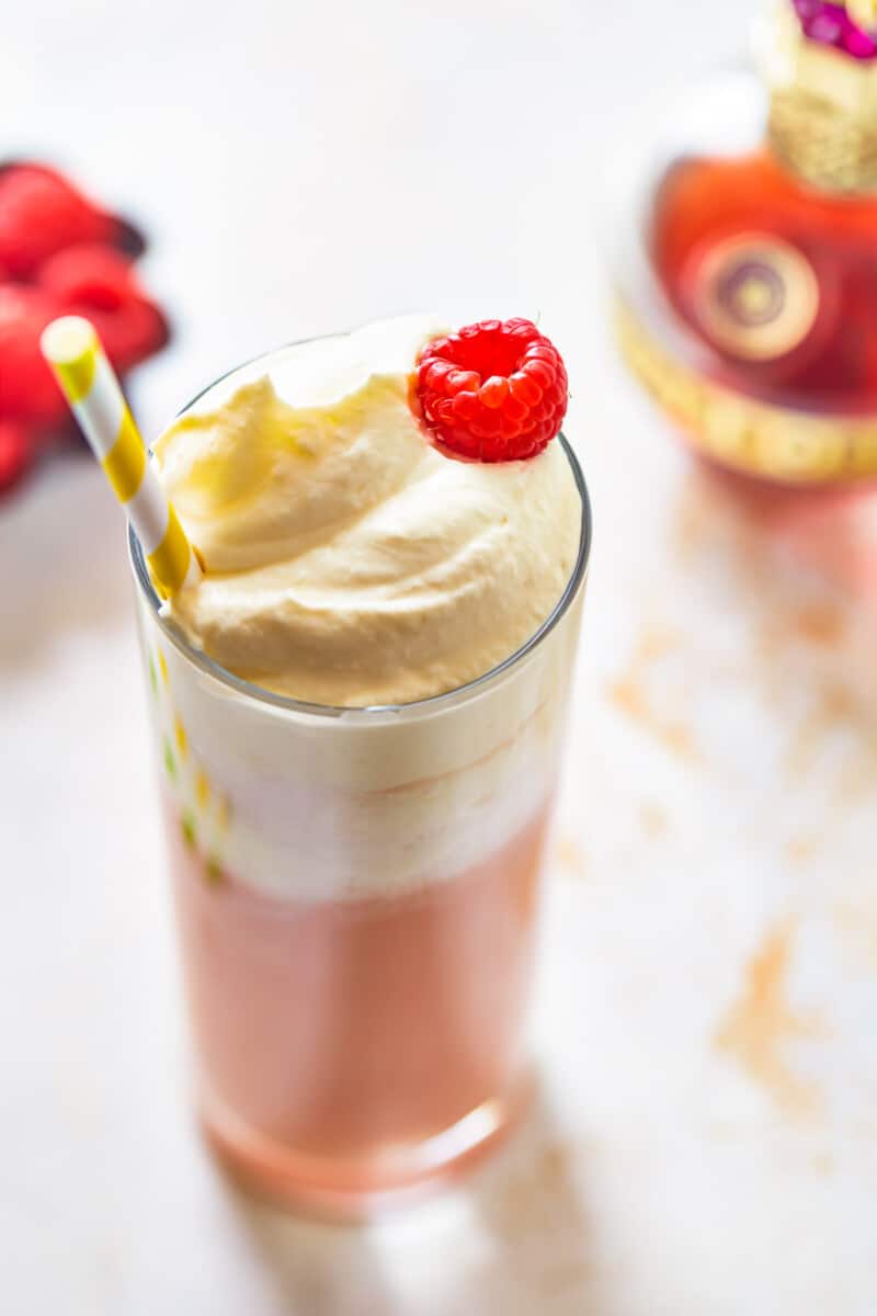 three-quarters view of raspberry italian cream soda in a tall glass topped with whipped cream, a raspberry, and a straw.