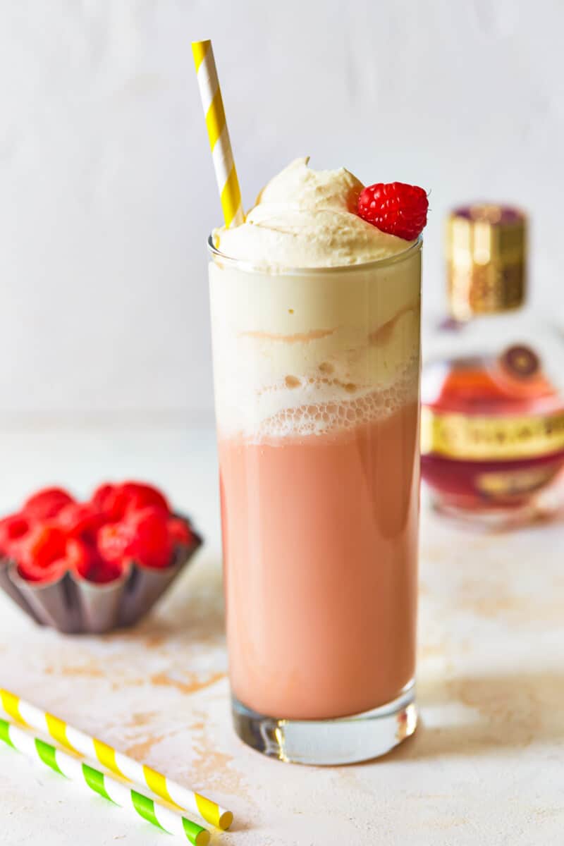 side view of raspberry italian cream soda in a tall glass topped with whipped cream, a raspberry, and a straw.