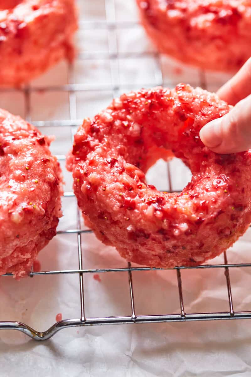 close up three-quarters view of a hand lifting a strawberry donut from a wire cooling rack.