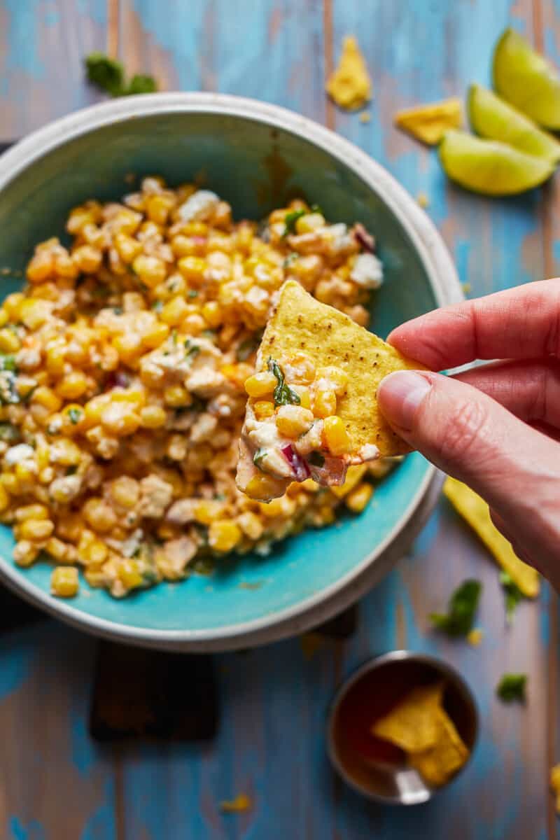 This Mexican Street Corn recipe is a healthy version of a restaurant favorite. I took the corn off the cob, switched out some ingredients (no mayo or sour cream), and kept the same delicious flavors to create this healthy Mexican Street Corn Salad recipe that works as an awesome appetizer, side dish, or snack! dipping chip into bowl of healthy mexican street corn dip