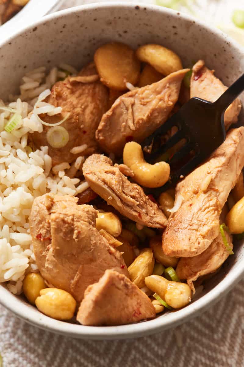 close up view of crockpot cashew chicken in a tan bowl with rice and a fork.