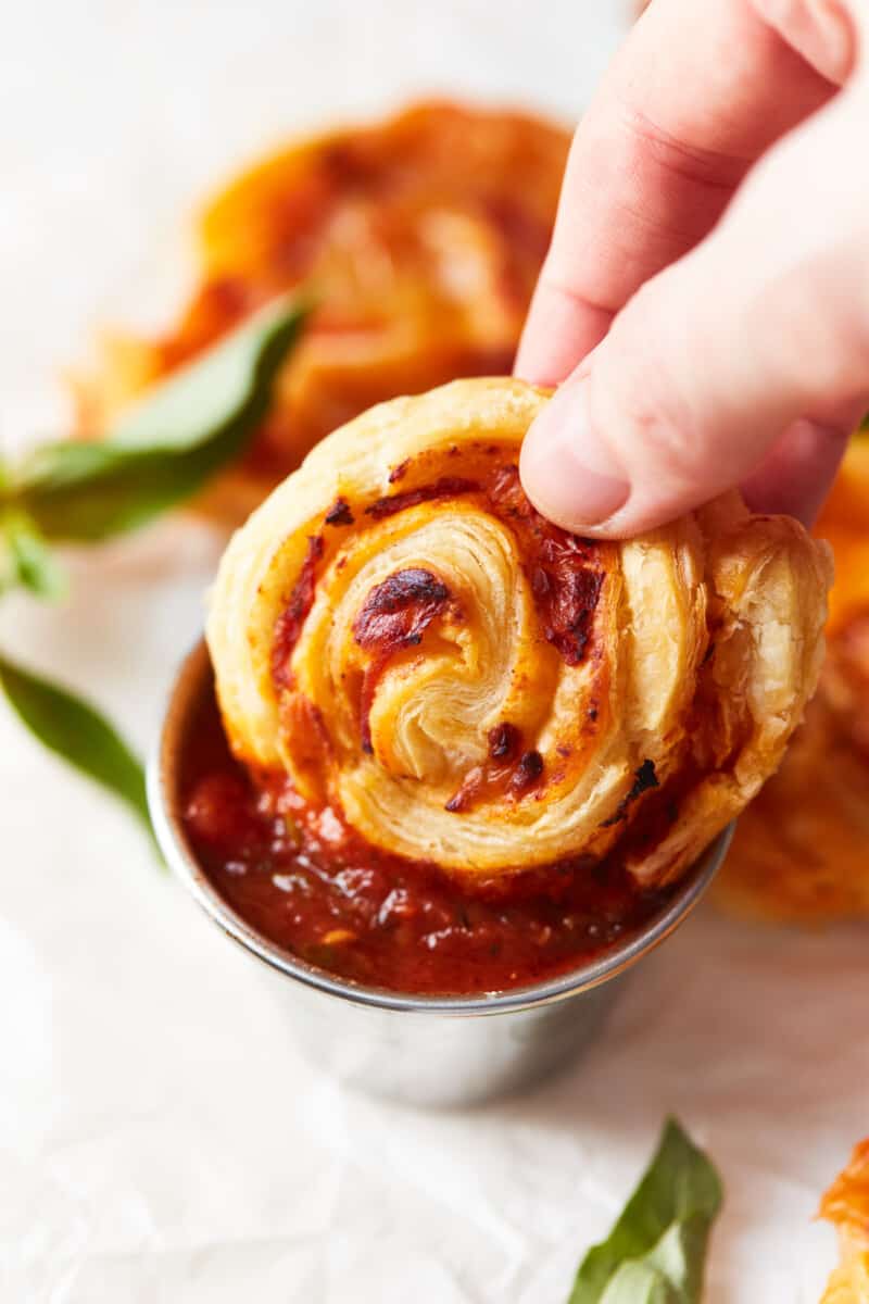 a hand dipping a pepperoni pizza roll into a small metal cup full of marinara sauce.