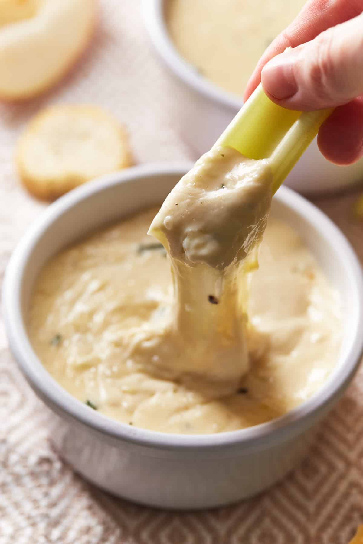 a hand pulling a piece of celery out of white wine fondue in a white bowl, creating a cheese pull.