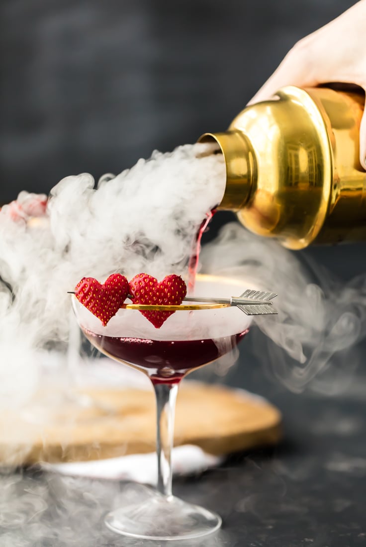 pouring a pomegranate martini out of a gold cocktail shaker into a glass with dry ice smoke
