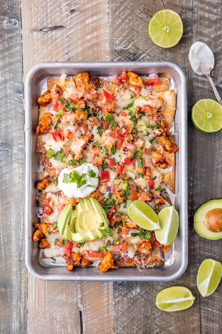 Chicken Nachos are great for feeding a crowd! This Tequila Lime Baked Chicken Nachos recipe is just AMAZING! These easy sheet pan nachos are so delicious, and perfect for any occasion. a sheet pan filled with chicken nachos on a wooden table surface