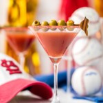 dirty redbird martini in a glass garnished with olives surrounded by baseballs and a saint louis cardinals hat