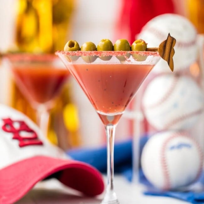 dirty redbird martini in a glass garnished with olives surrounded by baseballs and a saint louis cardinals hat