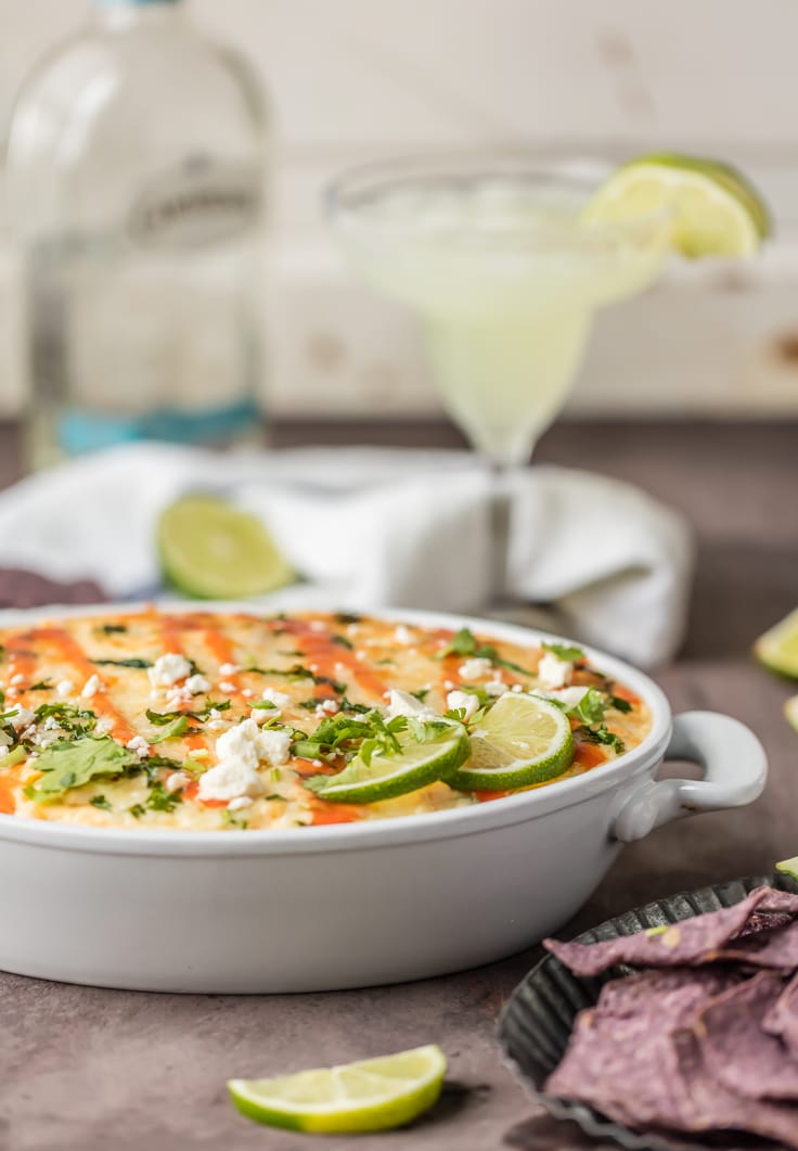 Side view of elote dip in a white baking dish topped with hot sauce, cheese, cilantro, and lime slices.