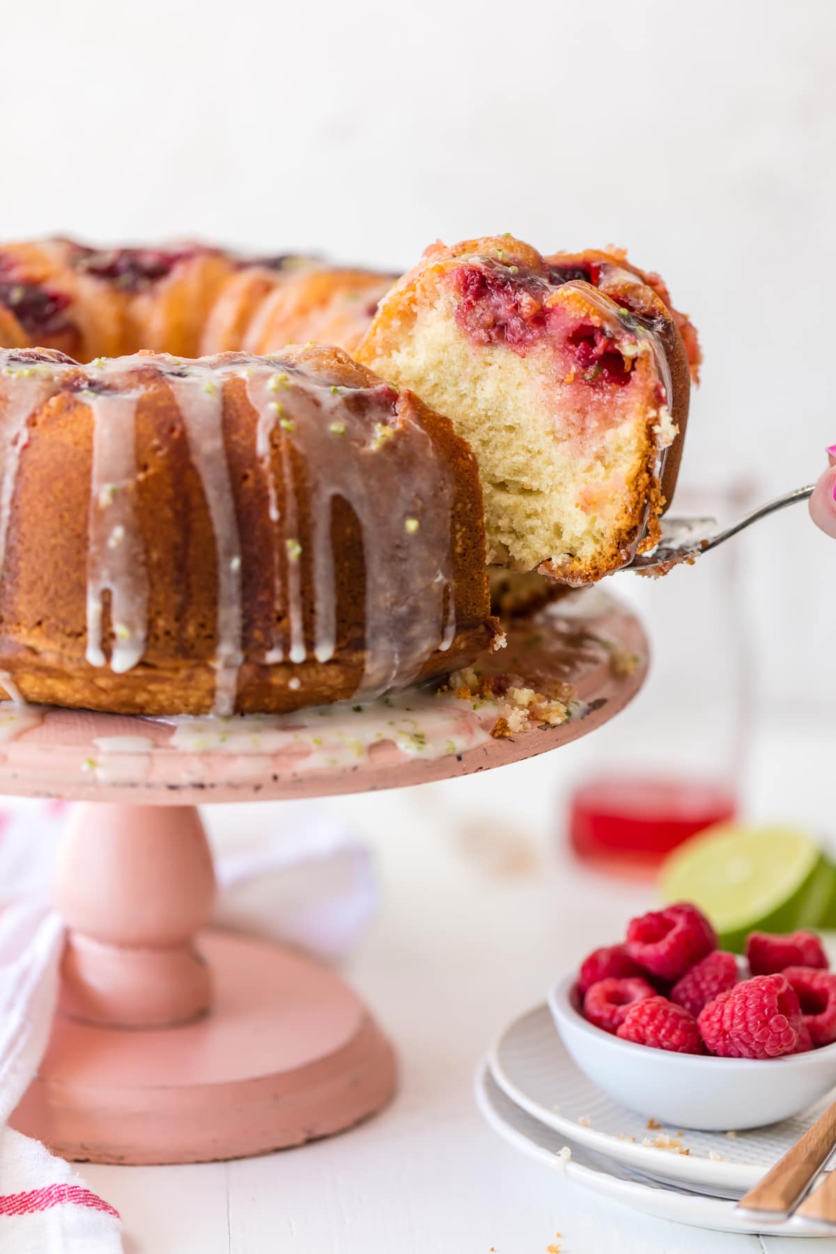 A raspberry cake on a pink cake stand next to a bowl of raspberries