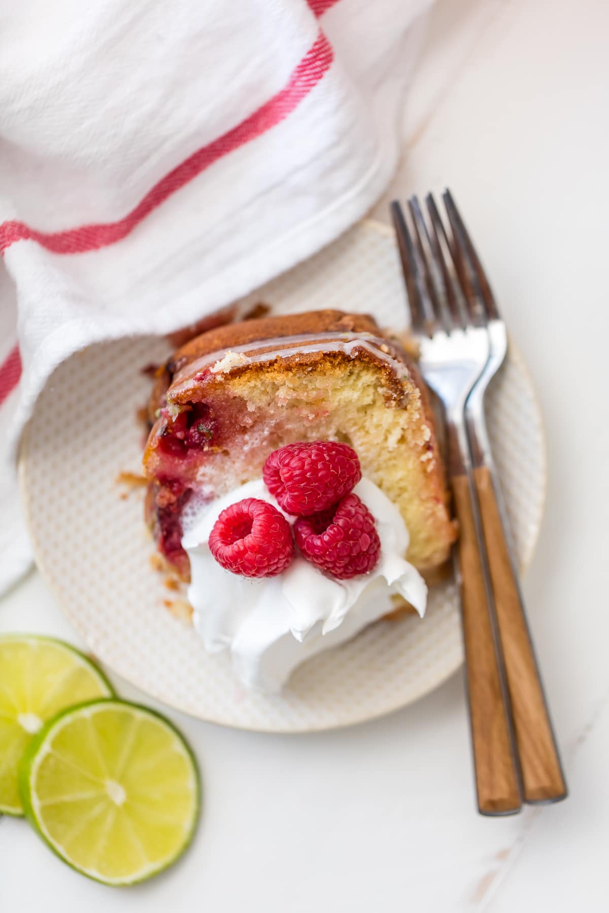 A slice of raspberry cake topped with cream and raspberries, on a white plate with two forks