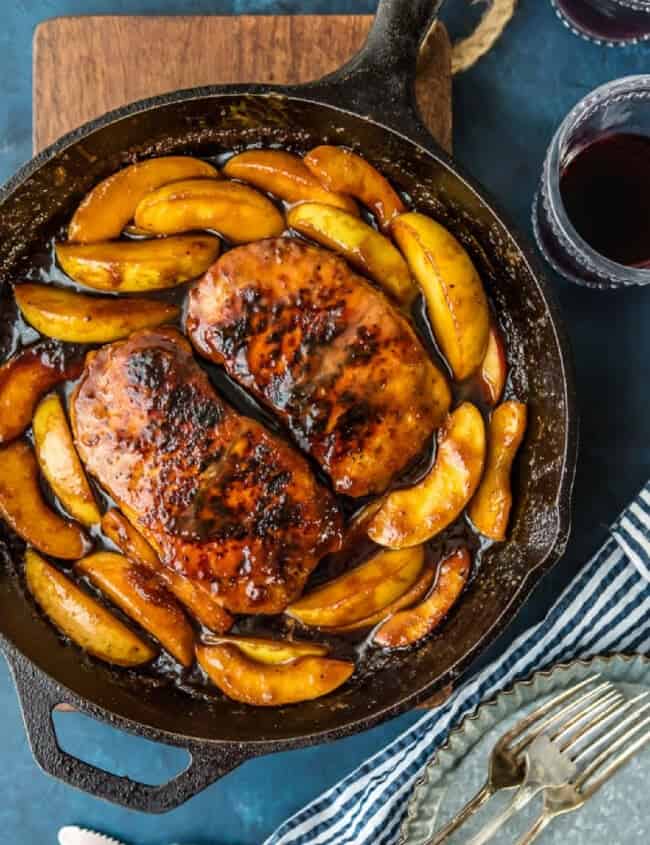 A cast iron skillet filled with pork chops and apples, accompanied by a glass of wine.
