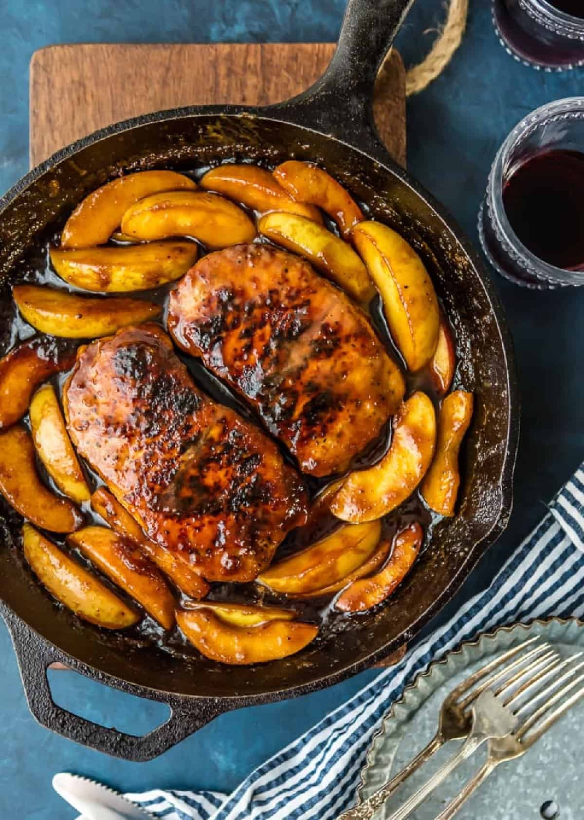 A cast iron skillet filled with pork chops and apples, accompanied by a glass of wine.