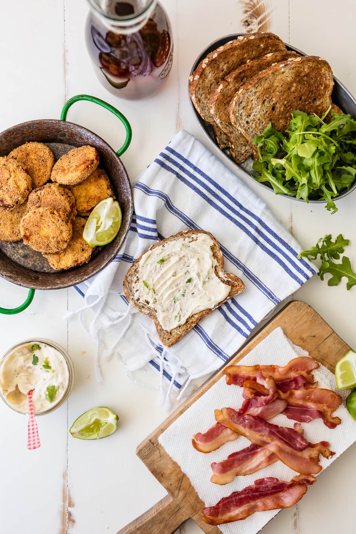 Skillet with fried tomatoes, plate of toasted bread, cutting board with bacon, and a piece of bread with mayo on a dish towel