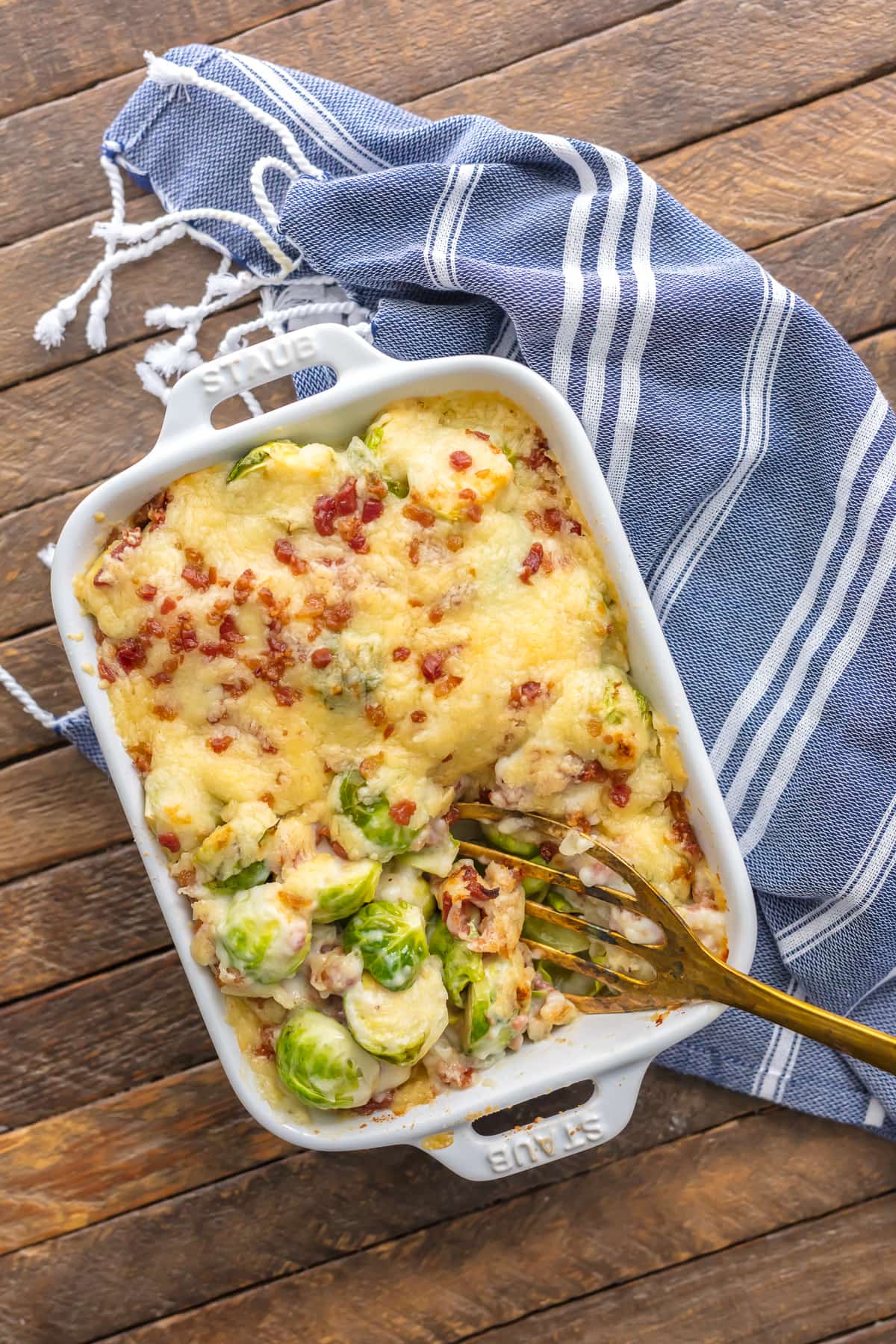 Bacon Brussels Sprouts Gratin in a white baking dish, sitting on a blue and white striped dish towel
