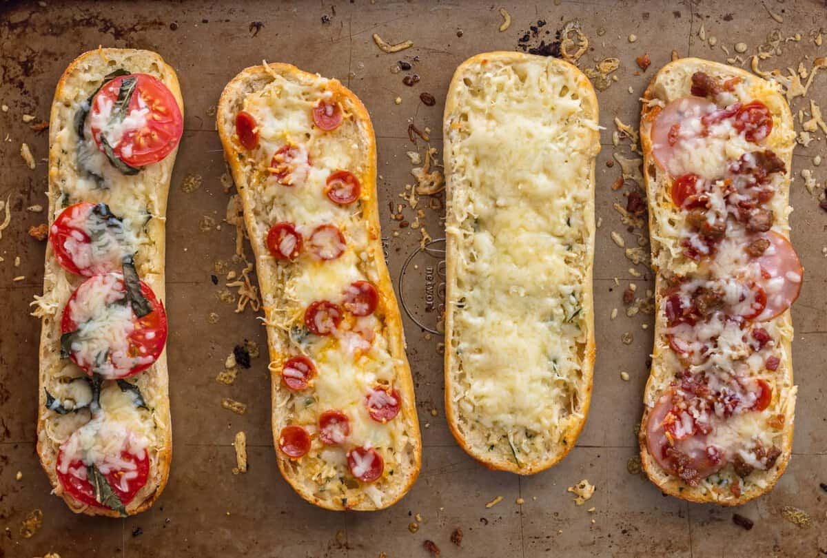 FRENCH BREAD PIZZA BITES are the easiest and tastiest cheesy appetizer! These mini french bread pizzas are loaded with garlic butter, cheese, and all of your favorite toppings. They make a great appetizer (the perfect party food), but we may just eat them as an entire meal! Four french bread pizza on a baking sheet: one topped with meat, one with cheese, one with pepperonis, one with tomatoes and basil