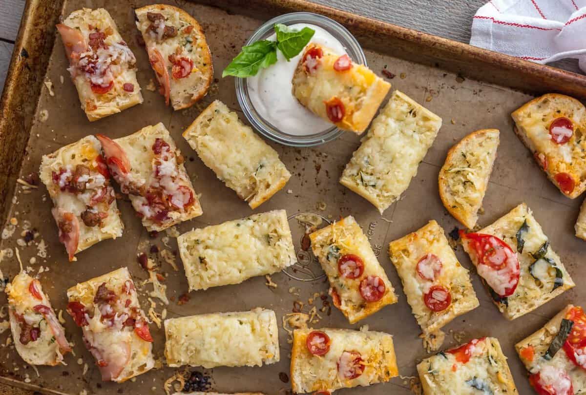 FRENCH BREAD PIZZA BITES are the easiest and tastiest cheesy appetizer! These mini french bread pizzas are loaded with garlic butter, cheese, and all of your favorite toppings. They make a great appetizer (the perfect party food), but we may just eat them as an entire meal! Garlic Bread Pizza Bites on a baking sheet with one piece in a bowl of ranch dressing.