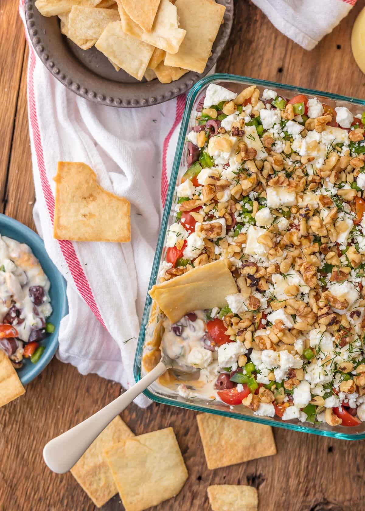 Greek 7 Layer Dip recipe in a baking dish sitting on a wooden table, next to a bowl of pita chips