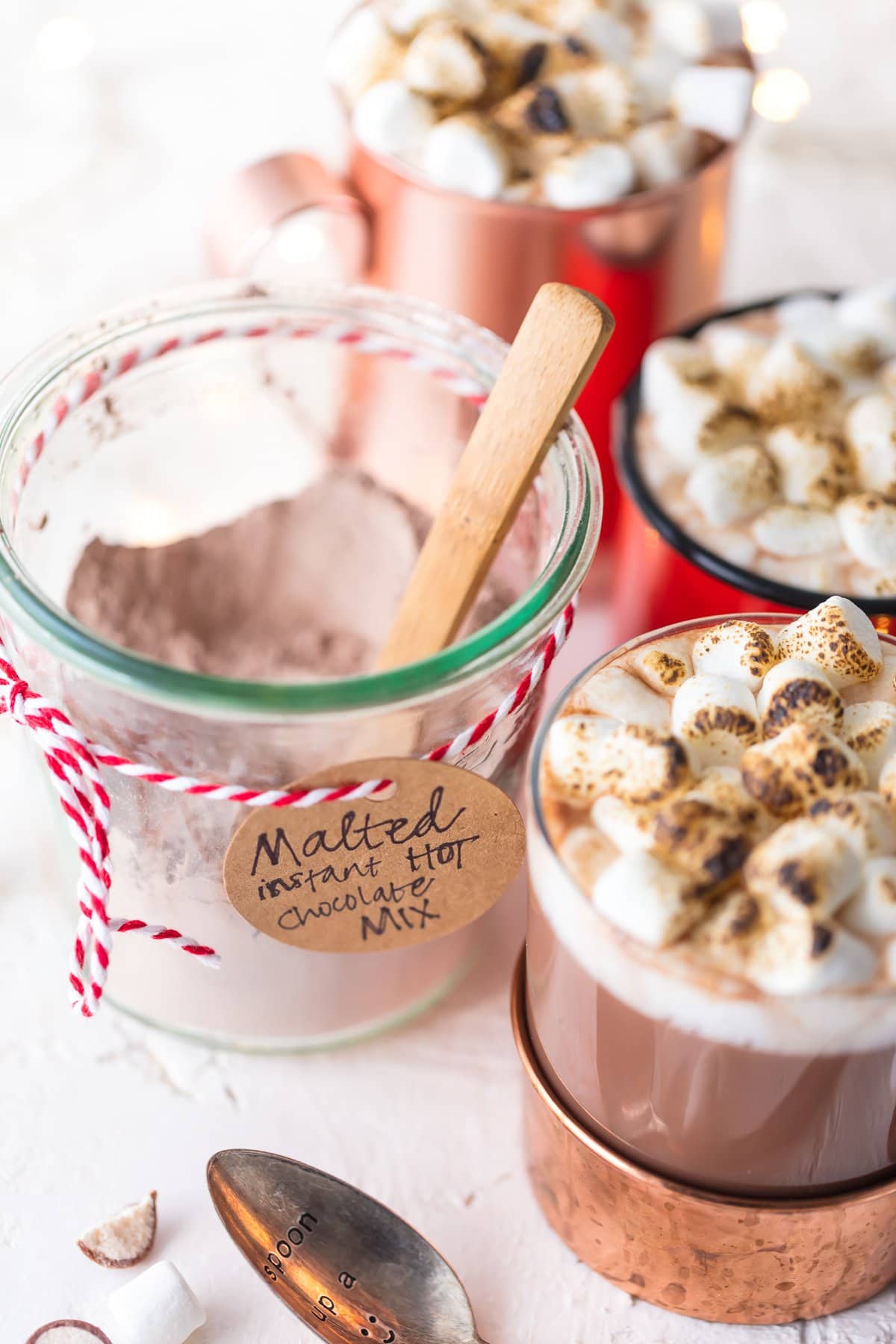 A jar of homemade hot chocolate mix next to three mugs of hot cocoa topped with marshmallows