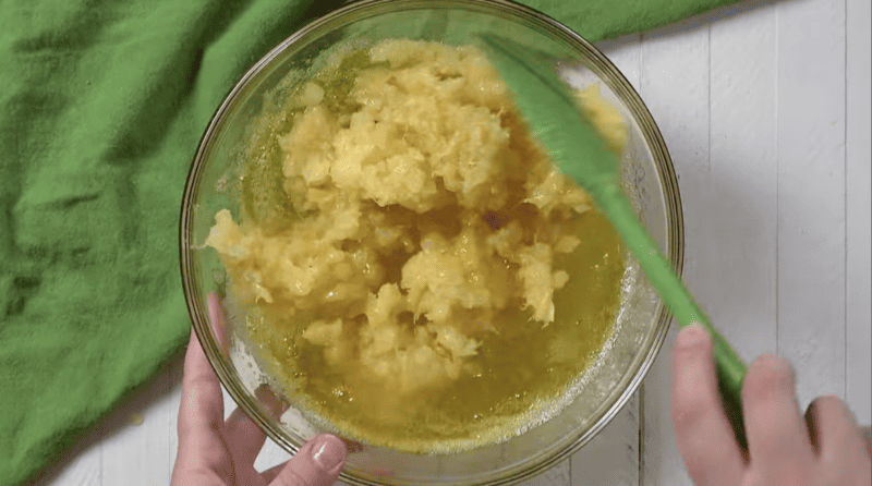 crushed pineapple folded into yellow liquid in a glass bowl with a green rubber spatula.