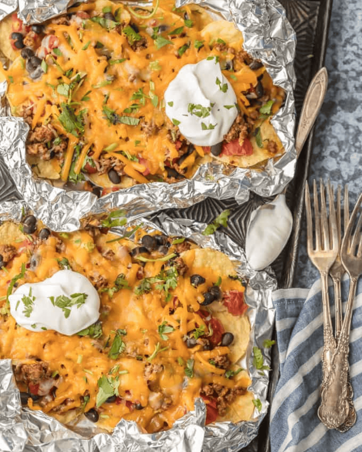 BEST Nachos Recipe - These foil packet nachos are loaded with beef, tomatoes, green chiles cheese, & more! I love this easy foil packet recipe, in the oven or on the grill. Foil packets with nachos garnished with sour cream.