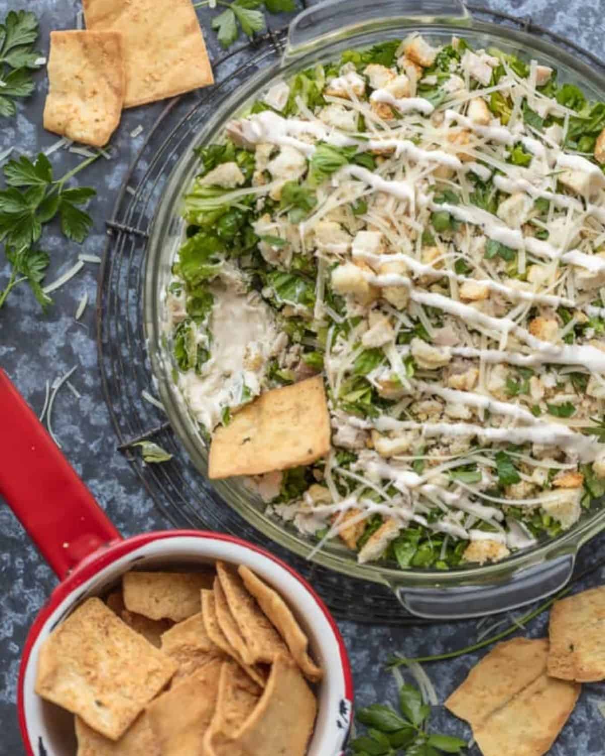This Caesar Salad Cream Cheese Chicken Dip recipe is the perfect mix of chicken caesar salad and cream cheese dip. The perfect appetizer for parties, serve it with pita chips or veggies! Chopped chicken Caesar salad in a glass bowl with pita chips.