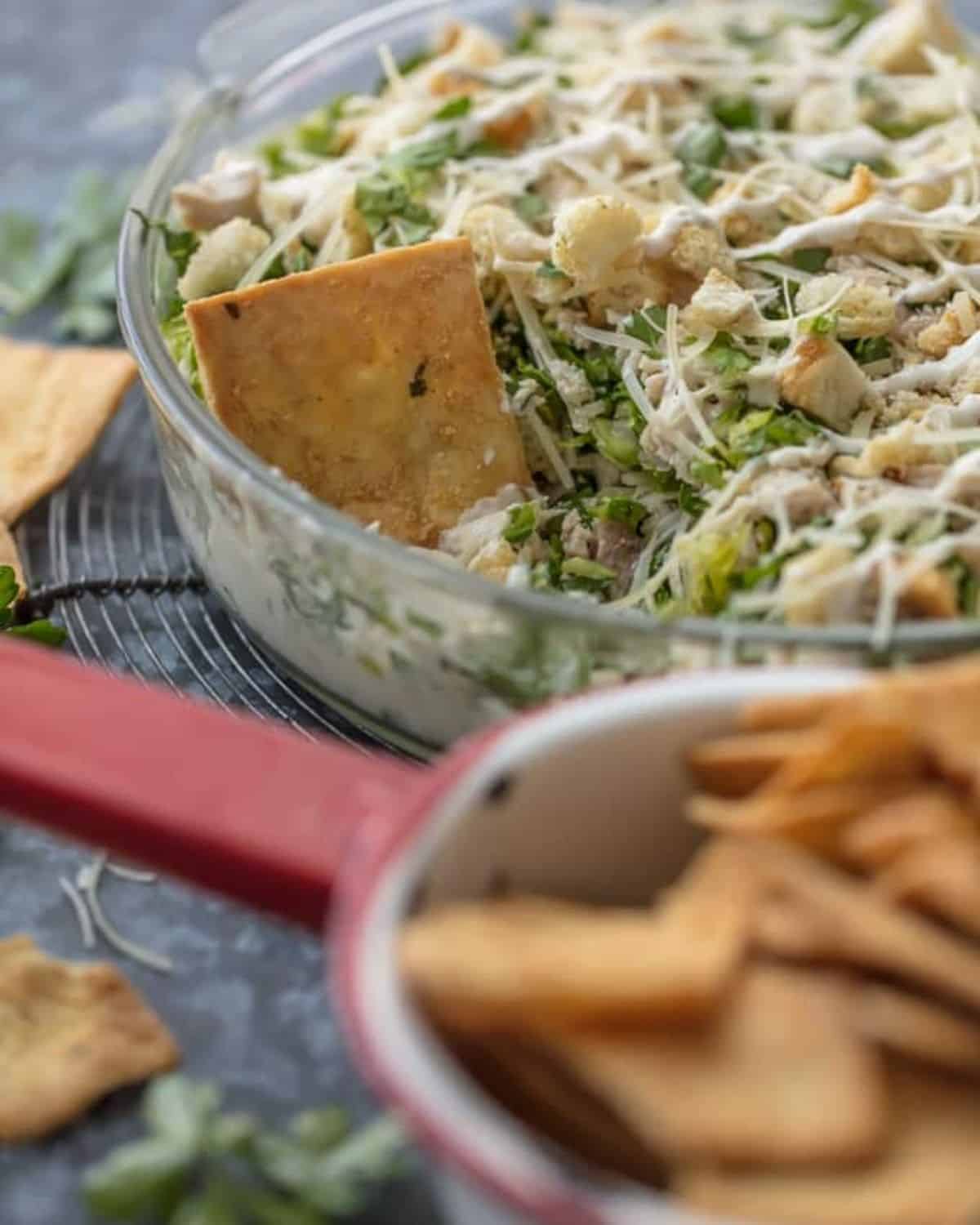 This Caesar Salad Cream Cheese Chicken Dip recipe is the perfect mix of chicken caesar salad and cream cheese dip. The perfect appetizer for parties, serve it with pita chips or veggies! Chopped chicken Caesar dip in a glass bowl with a pita chip in it.