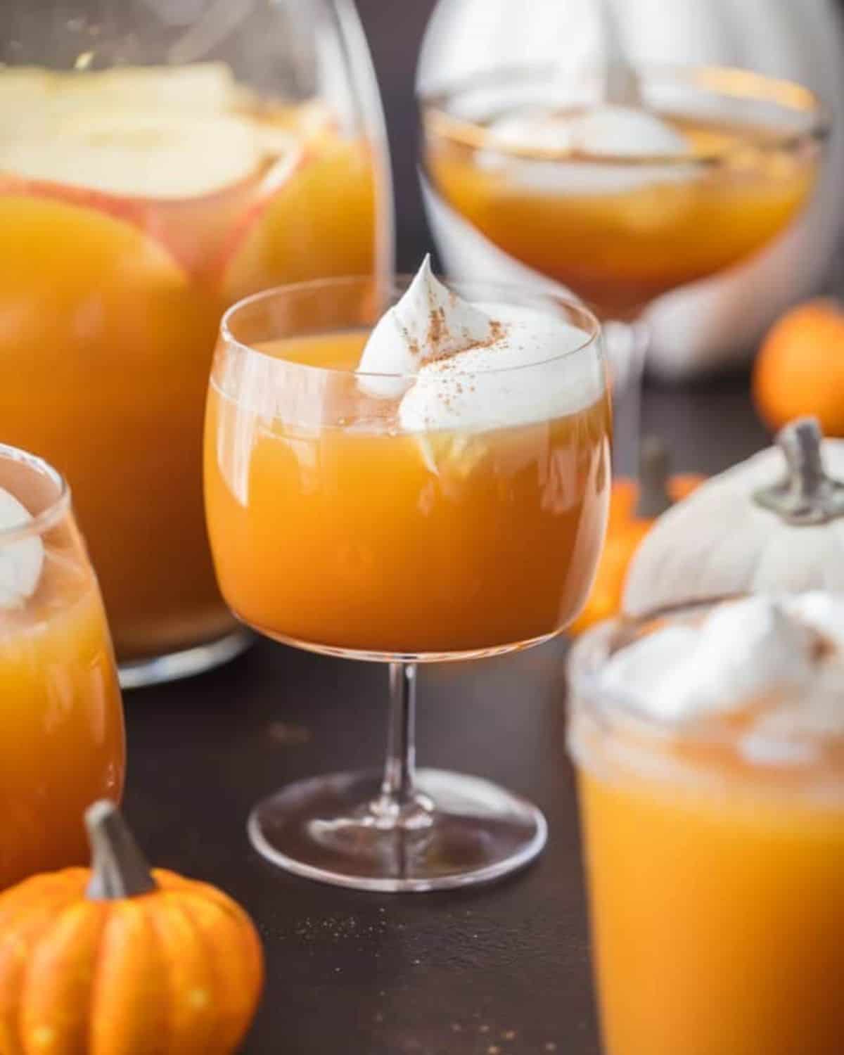 This Pumpkin Punch recipe is the perfect Thanksgiving or Halloween Punch idea. A fun & tasty Pumpkin Cocktail inspired by pumpkin pie! Pumpkin Pie Punch in a glass with a few glasses of punch around it and a mini pumpkin.