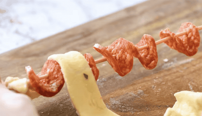 Pepperoni threaded onto a skewer with pizza dough being wrapped around it.