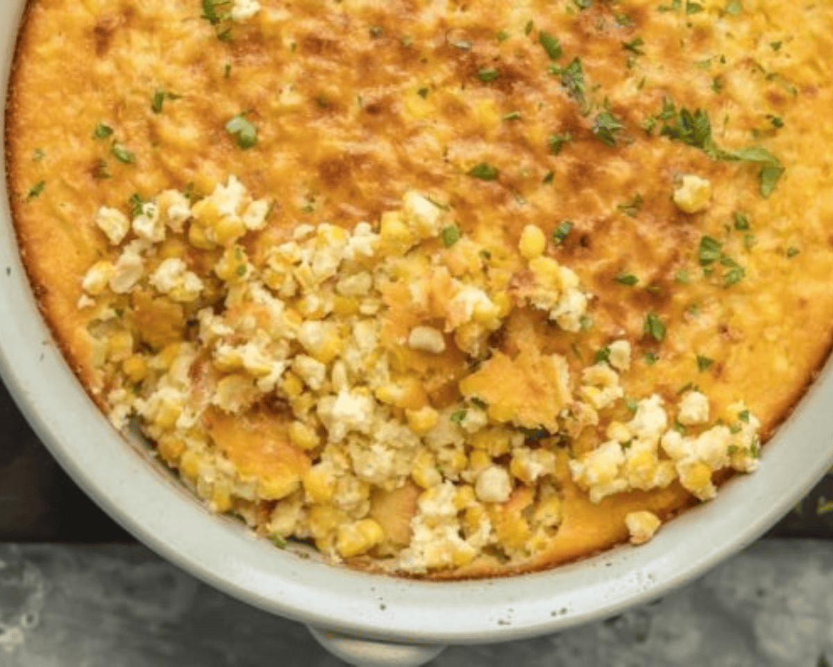 Close-up of baked corn pudding in a white casserole dish with a portion scooped out, showing creamy corn filling beneath the golden top