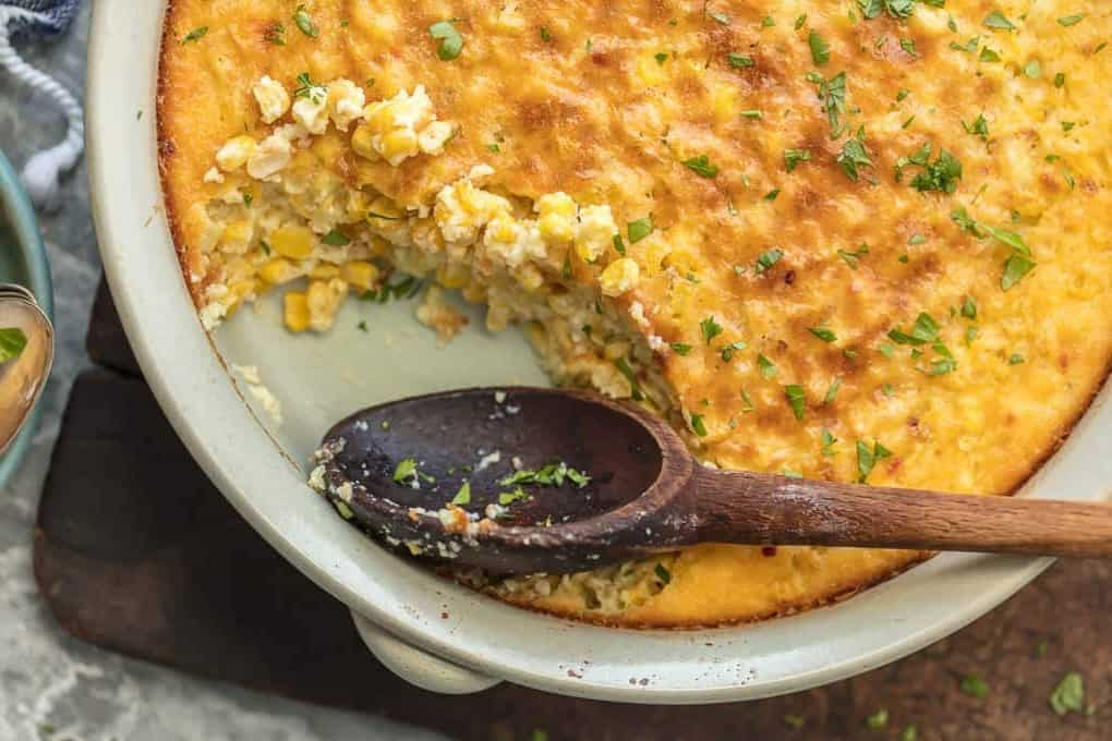 Baked corn pudding with a golden crust in a white casserole dish, a wooden spoon resting inside after scooping out a serving