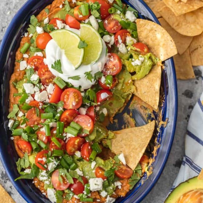 This Layered Bean Dip Recipe is one of our favorite ways to tailgate! Guacamole Bean Dip has layers of guacamole, spicy refried beans, tomatoes, onions, and cheese. This Mexican Bean Dip is SO delicious and made in minutes! It doesn't get better than Layered Bean Dip for any and every celebration! Learn how to make bean dip with this recipe.