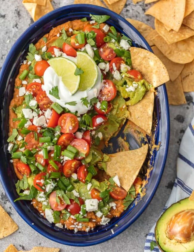 This Layered Bean Dip Recipe is one of our favorite ways to tailgate! Guacamole Bean Dip has layers of guacamole, spicy refried beans, tomatoes, onions, and cheese. This Mexican Bean Dip is SO delicious and made in minutes! It doesn't get better than Layered Bean Dip for any and every celebration! Learn how to make bean dip with this recipe.