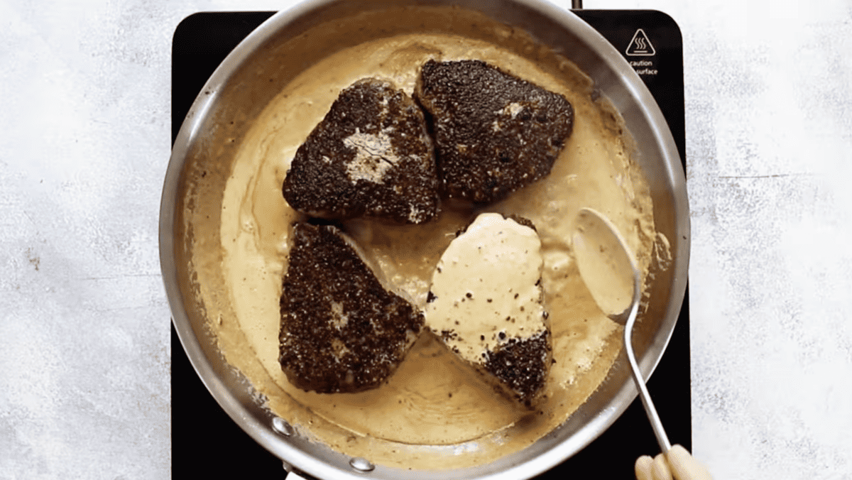 A person pouring a brandy cream sauce over a peppercorn steak in a pan.