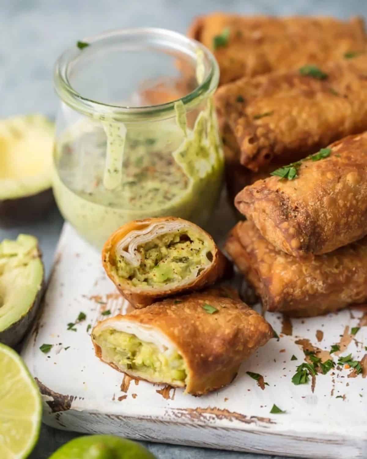 This Avocado Egg Rolls Recipe is SO delicious and addicting. This fun deep fried egg roll recipe is stuffed with the best guacamole ever, and paired with a green sauce you have to taste to believe. Every party needs these GUACAMOLE EGG ROLLS! If you've ever wondered how to make egg rolls, it's easier than you think! Anyone can make this Easy Egg Roll Recipe and everyone will love them. Avocado spring rolls with a glass jar of Peruvian green sauce.