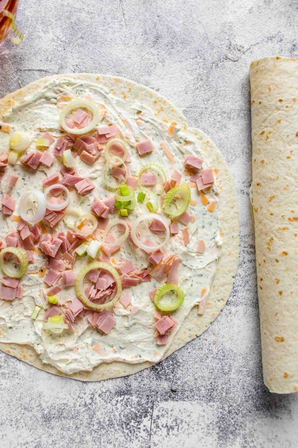 tortilla laid flat with cream cheese, ham, cheese, ranch, and green onions spread out on top.