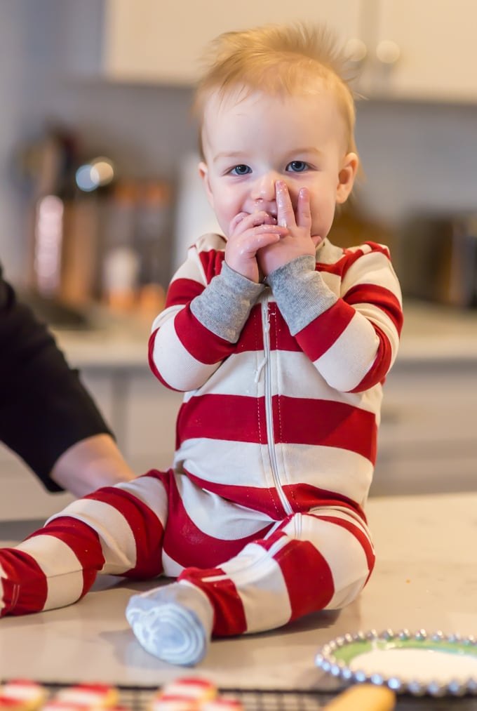 blond haired baby boy in his red and white striped Christmas pajamas as he licks sugar cookie frosting from his fingers