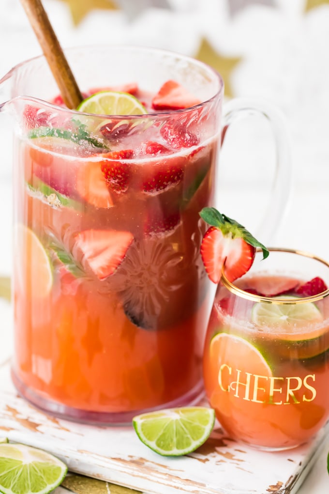 a pitcher of sparkling margarita punch filled with slices of fresh limes and strawberries. a glass of the punch sits next to the pitcher,