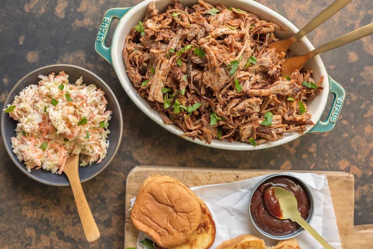 Serving dish filled with BBQ pulled pork, a bowl of coleslaw, a bowl of BBQ sauce, and a bun.