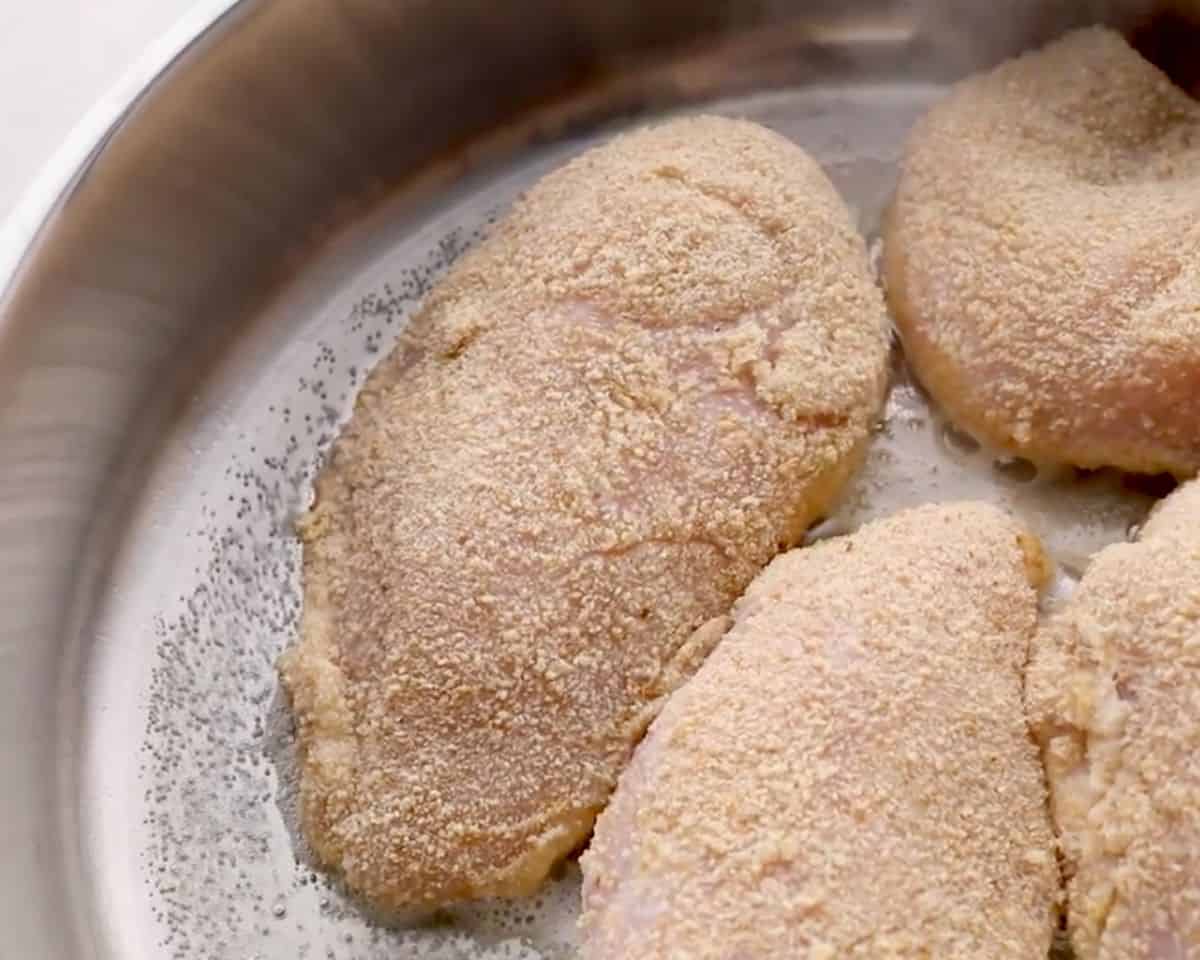 Breaded chicken breasts cooking in a stainless steel skillet before browning