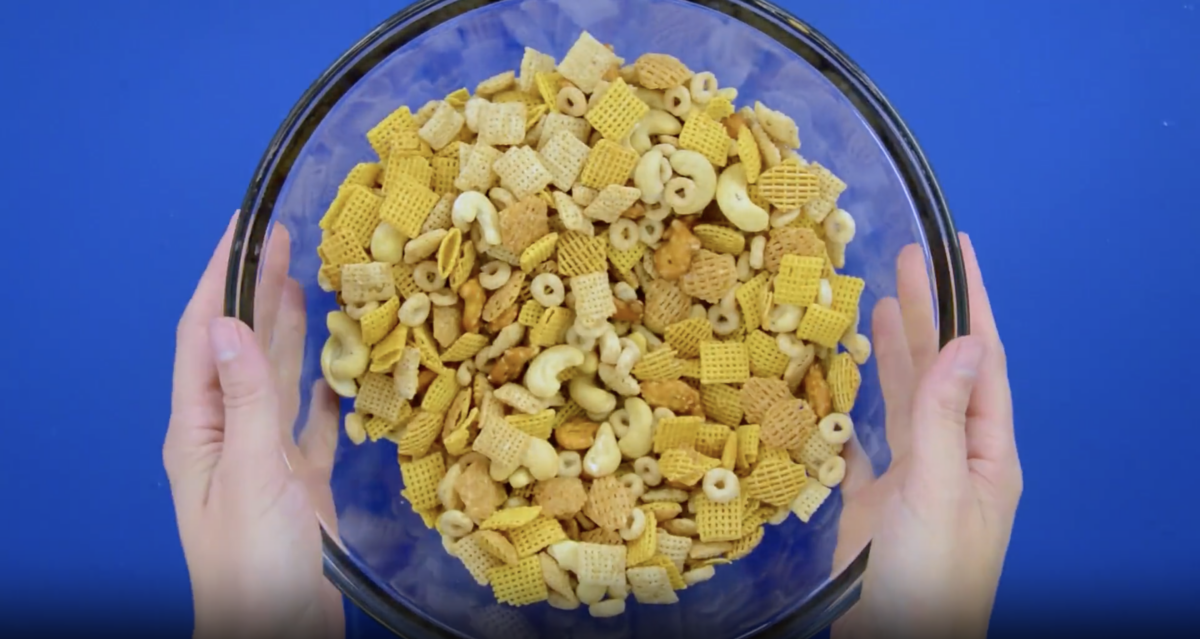 A person holding a bowl of chex party mix on a blue background.