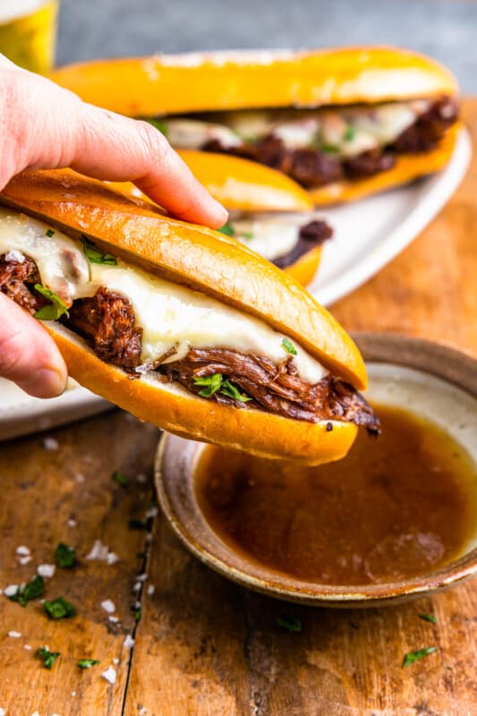 side view of a hand dipping a crockpot french dip sandwich into a side of au jus.