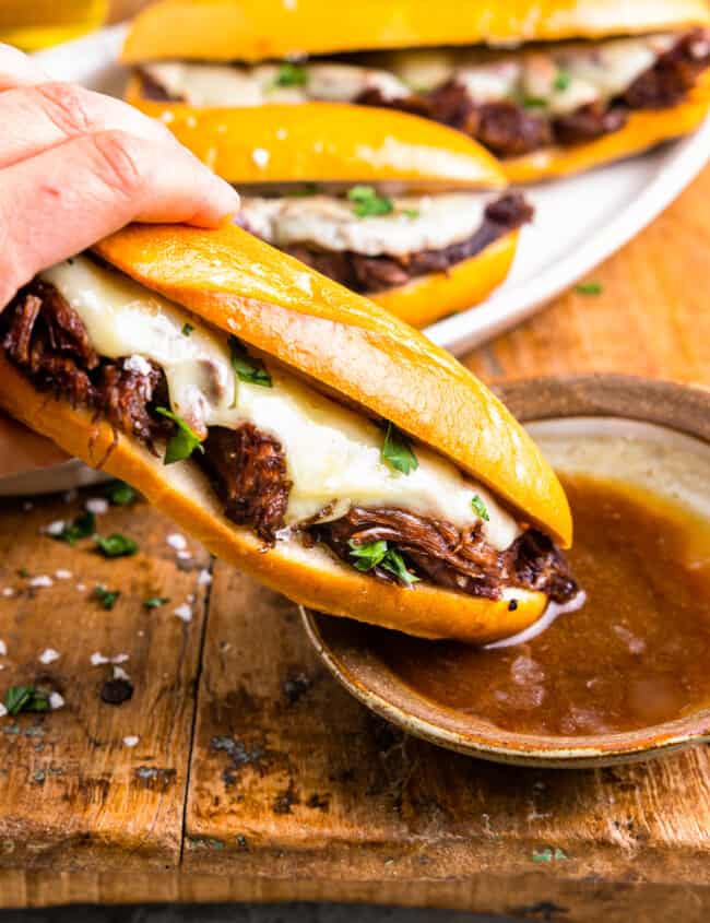 side view of a hand dipping a crockpot french dip sandwich into a side of au jus.